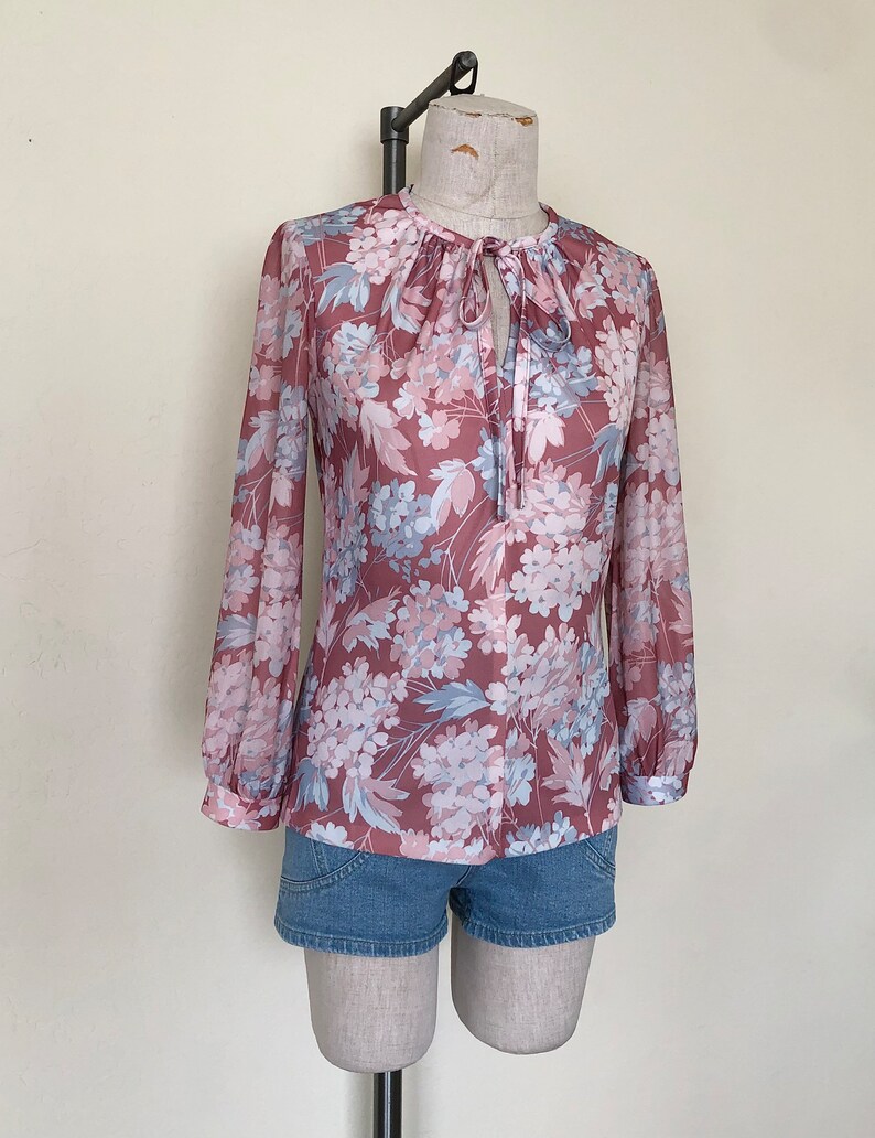 May include: A long-sleeved blouse with a floral print in shades of pink, blue, and white. The blouse has a tie at the neck and a loose fit.