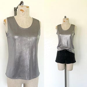 May include: A silver metallic tank top with a scoop neck is displayed on a mannequin. The sleeveless top has a shiny, reflective surface. The mannequin is on a stand against a white background. The tank top is shown in two different views.
