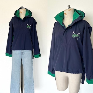 Puede incluir: Chaqueta anorak azul marino con capucha y puños forrados en verde. La chaqueta presenta un diseño bordado en verde en el pecho. El anorak se muestra en un maniquí y se combina con vaqueros azul claro.