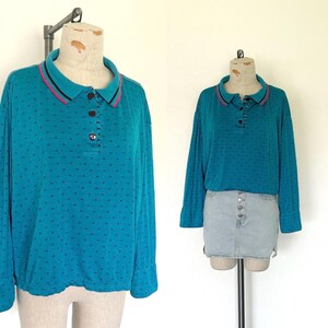 May include: Teal long-sleeved shirt with a polo collar. The collar has a pink and black stripe detail. The shirt has a button-up placket and small dark dots. The shirt is displayed on a mannequin.