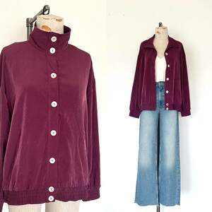 May include: A burgundy jacket with a high collar and white buttons. The jacket has long sleeves and a ribbed hem. The jacket is displayed on a mannequin. The image also shows a second burgundy jacket with a white top and blue jeans.