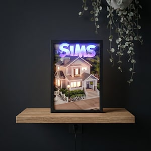 May include: A framed, illuminated diorama of a house, with the text "The Sims" at the top. The house is a two-storey structure with a porch, lit windows, and a small yard with bicycles. The frame is black, and the scene is set against a dark background.