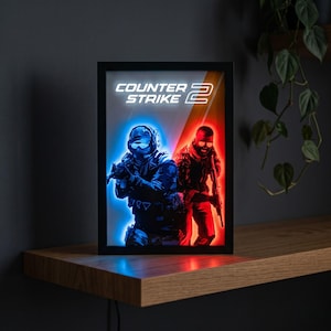 Puede incluir: Obra de arte enmarcada e iluminada que representa a dos soldados en un videojuego, iluminados en azul y rojo. Las palabras "Counter Strike 2" se muestran en la parte superior. La obra está sobre un fondo oscuro.