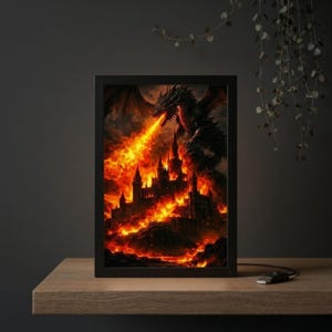 May include: A framed print depicting a black dragon breathing fire onto a castle engulfed in flames. The artwork features a dark color palette with fiery orange and red hues, creating a dramatic fantasy scene. The print is in a black frame.