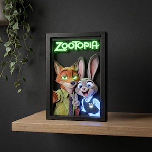 May include: A framed Zootopia art piece featuring Nick Wilde and Judy Hopps. The artwork has a 3D effect, with the characters in the foreground. The Zootopia logo is illuminated in green. The frame is black and sits on a wooden shelf.