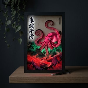 May include: Framed art piece featuring a vibrant pink octopus over a red and green landscape with Japanese text. The artwork is illuminated, creating a striking visual effect. The frame is black, and the piece is displayed on a wooden surface.