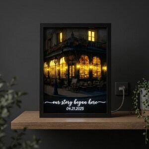 Könnte beinhalten: Gerahmter Druck mit einer nächtlichen Szene eines europäischen Cafés, beleuchtet von warmen Lichtern. Der Text "our story began here" und das Datum 04.21.2025 sind in eleganter Schrift unten dargestellt. Der Rahmen ist schwarz.