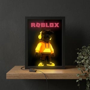 May include: Illuminated framed artwork of a Roblox character wearing a yellow hoodie, sunglasses, and headphones. The pink Roblox logo is above the character. The artwork is set against a black background, with a wooden shelf and a plant in the background.