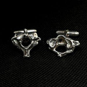 May include: A pair of silver-toned cufflinks, each designed in the shape of a vertebra. The cufflinks have a detailed, anatomical appearance, with a post and toggle closure. The background is a dark, textured surface.