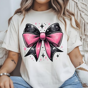 Könnte beinhalten: Cremefarbenes T-Shirt mit einer großen Grafik einer rosa und schwarzen Schleife mit Sternennacht-Design. Die Schleife ist mit rosa und schwarzen Sternen verziert, und das Shirt hat kurze Ärmel.