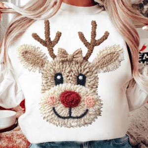 Puede incluir: Sudadera blanca de manga larga con un diseño de cara de reno. El reno tiene cuernos marrones, una nariz roja y una boca sonriente. El diseño está hecho de un material texturizado y difuso, ideal para la temporada navideña.