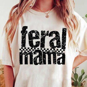 May include: Cream-colored t-shirt with the words "feral mama" in distressed black font. The word "feral" is above "mama", with a black and white checkered pattern between them. The shirt is a casual style.