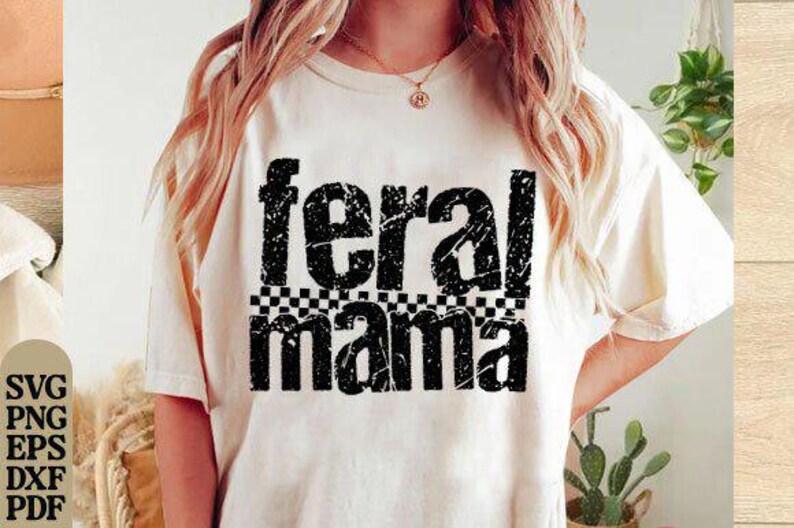 May include: Cream-colored t-shirt with the words "feral mama" in distressed black font. The word "feral" is above "mama", with a black and white checkered pattern between them. The shirt is a casual style.