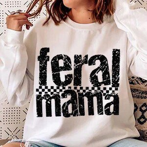 May include: White sweatshirt with the text "feral mama" in a distressed black font. The text is above a black and white checkered pattern. The sweatshirt has long sleeves.