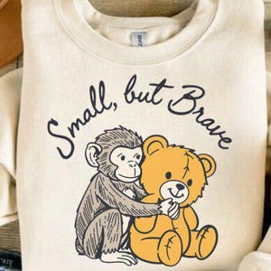 Op de afbeelding: Crèmekleurige sweatshirt met een cartoonillustratie van een aap die een teddybeer omhelst. De tekst "Small, but Brave" is in een handschriftstijl boven de illustratie gebogen. Het ontwerp is gecentreerd op de voorkant van de sweatshirt.