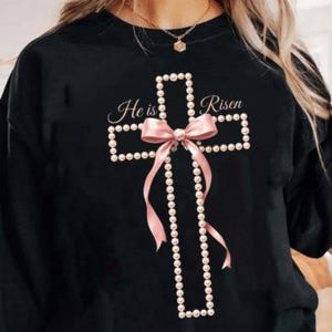 Könnte beinhalten: Schwarzes Sweatshirt mit einem Kreuzdesign aus weißen Perlen, einer rosa Schleife und Bändern. Die Worte "He is Risen" sind in Schrift über dem Kreuz geschrieben. Eine Halskette ist sichtbar.