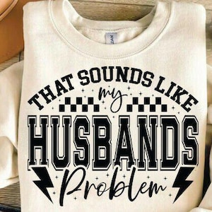 May include: Cream-colored sweatshirt with the text "THAT SOUNDS LIKE my HUSBANDS Problem" in black. The design includes a checkered pattern and lightning bolt graphics, perfect for casual wear.