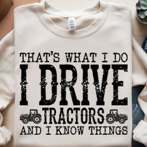 May include: Cream-colored sweatshirt with the text "THAT'S WHAT I DO I DRIVE TRACTORS AND I KNOW THINGS" in black. Two tractor graphics are below the text. The sweatshirt is folded on a light-colored surface.