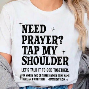 Pode incluir: Camiseta branca com o texto preto "NEED PRAYER? TAP MY SHOULDER". A camiseta também inclui a frase "LET'S TALK IT TO GOD TOGETHER" e um versículo bíblico. É uma peça de roupa casual.