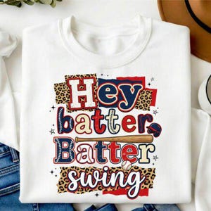 Può includere: Felpa bianca con la scritta "Hey Batter Batter Swing" in rosso, blu e beige. Il design include una mazza da baseball, una palla da baseball e dettagli con stampa leopardata. Un capo d'abbigliamento a tema baseball.