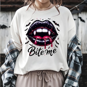 May include: Cream-colored t-shirt featuring a graphic of black lips with fangs, dripping red blood, and the text "Bite me." The design includes bats and stars, creating a spooky Halloween theme.