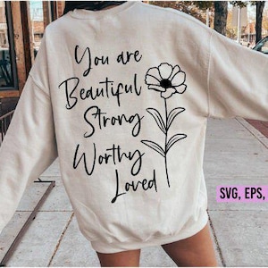 May include: Cream-colored sweatshirt with the words "You are Beautiful Strong Worthy Loved" in black script, accompanied by a black floral design. The sweatshirt also has the text "SVG, EPS, DXF, PNG" in a pink rectangle.