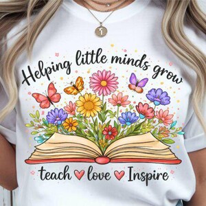 May include: White t-shirt featuring a colorful illustration of an open book with flowers and butterflies. The text "Helping little minds grow teach love inspire" is written above and below the illustration. The design is in a watercolor style.