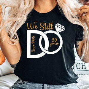 Pode incluir: Camiseta preta com texto dourado e branco celebrando um aniversário de 20 anos. O design apresenta um gráfico de anel de diamante e as palavras "We Still Do Since 2005" em uma fonte elegante.