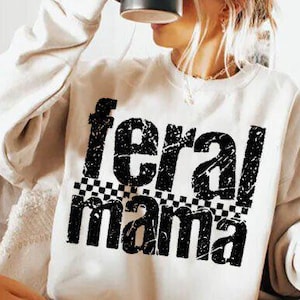 May include: Cream-colored sweatshirt with the text "feral mama" in distressed black lettering. The word "feral" is above "mama", with a checkered pattern between the words. The sweatshirt is a casual style.