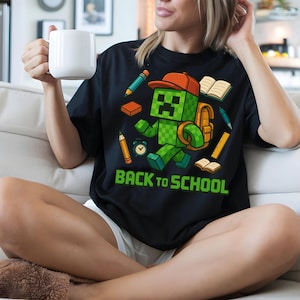 May include: Black t-shirt featuring a green pixelated character with an orange hat and backpack, surrounded by school supplies. The words "BACK TO SCHOOL" are printed in green below the character. The shirt is worn by a person.