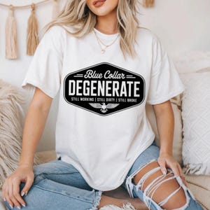 May include: Cream-colored t-shirt with a black and blue graphic. The graphic reads "Blue Collar Degenerate" with the phrases "Still Working | Still Dirty | Still Broke" below. A stylized eagle is at the bottom of the graphic.