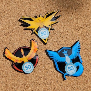 May include: Three enamel pins featuring Pokemon characters. The pins are yellow, orange, and blue. Each pin has a blue circle with the number 50 in white.