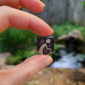 May include: A small, black and white plastic sign with a white stick figure of a person using a cane. The sign is held between two fingers.