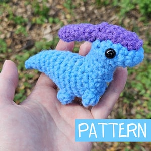 May include: A crocheted dinosaur toy with a blue body and purple crest. The dinosaur has black eyes and is being held in a hand. The word "PATTERN" is written in white on a blue background.
