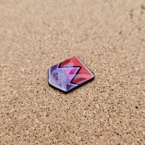 May include: A small, geometric resin pendant with a black border. The pendant features a vibrant design with red, purple, and white hues. The shape is a modified hexagon with a zig-zag pattern. The pendant is displayed on a textured, tan surface.
