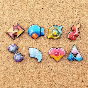 May include: Eight colorful enamel pins featuring different shapes and designs. The pins are arranged in two rows of four on a corkboard background.