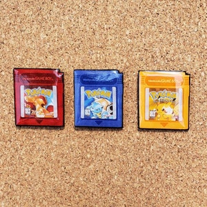 May include: Three Nintendo Game Boy cartridges featuring Pokemon characters. The cartridges are red, blue, and yellow. The red cartridge features Charizard, the blue cartridge features Blastoise, and the yellow cartridge features Pikachu.