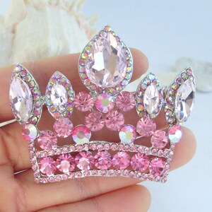 以下が含まれることがあります： シルバーカラーのメタルベースにピンクのラインストーンクラウンブローチ。王冠は、大きなティアドロップ型と丸いピンクのラインストーンが特徴で、ベースに沿って小さなピンクの石が付いています。ブローチは手で持っています。