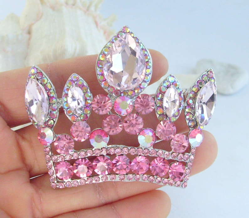 以下が含まれることがあります： シルバーカラーのメタルベースにピンクのラインストーンクラウンブローチ。王冠は、大きなティアドロップ型と丸いピンクのラインストーンが特徴で、ベースに沿って小さなピンクの石が付いています。ブローチは手で持っています。