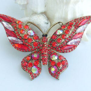 May include: A large, ornate butterfly brooch with a gold-tone metal frame. The wings are covered in small, round red rhinestones and larger, clear, and iridescent stones. The butterfly's body is black, and the antennae are gold.