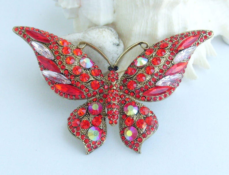 May include: A large, ornate butterfly brooch with a gold-tone metal frame. The wings are covered in small, round red rhinestones and larger, clear, and iridescent stones. The butterfly's body is black, and the antennae are gold.