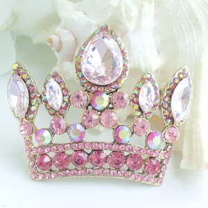 以下が含まれることがあります： ピンクと虹色のラインストーンで飾られた、金色の王冠ブローチ。王冠の中央には大きな洋梨型のピンク色の石があり、その周りを小さなピンクと虹色の石が囲んでいます。デザインには複数のアーチと、ベースにピンク色の石の列が含まれています。
