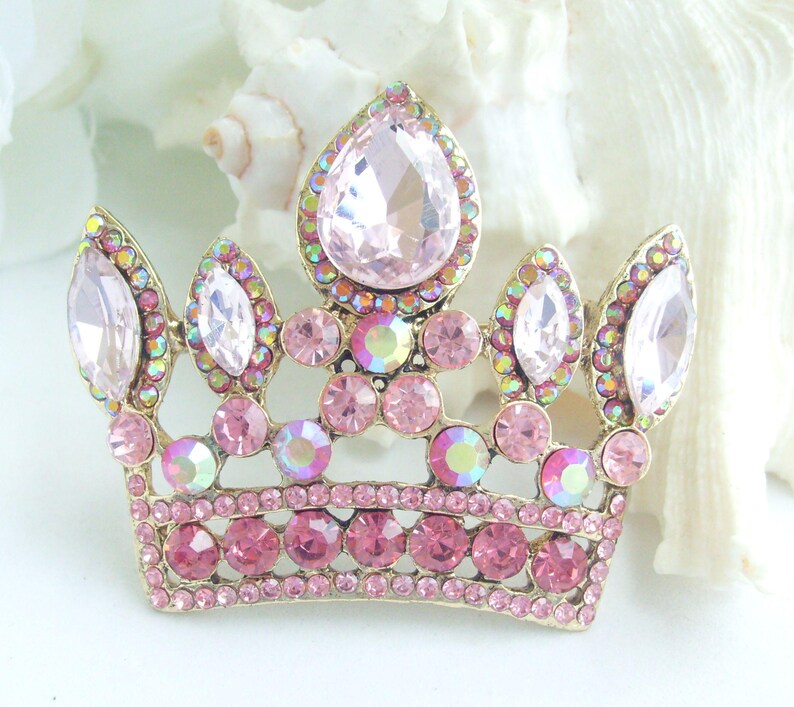 以下が含まれることがあります： ピンクと虹色のラインストーンで飾られた、金色の王冠ブローチ。王冠の中央には大きな洋梨型のピンク色の石があり、その周りを小さなピンクと虹色の石が囲んでいます。デザインには複数のアーチと、ベースにピンク色の石の列が含まれています。