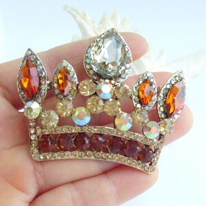 以下が含まれることがあります： 中央に涙型の石、オレンジ色と透明な宝石がいくつか付いた、金色の王冠ブローチ。 王冠の基部には濃い赤色の石が並んでいます。