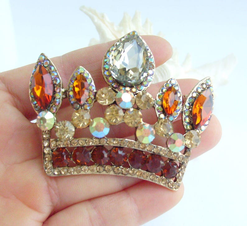 以下が含まれることがあります： 中央に涙型の石、オレンジ色と透明な宝石がいくつか付いた、金色の王冠ブローチ。 王冠の基部には濃い赤色の石が並んでいます。