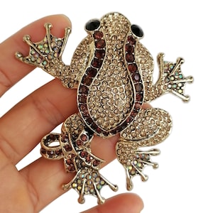 May include: A gold-toned frog brooch encrusted with rhinestones. The frog's body is covered in small, light-brown rhinestones, with a central stripe of darker, burgundy-colored stones. The eyes are black, and the feet have iridescent accents.