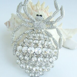 Może przedstawiać: Srebrna broszka w kształcie kraba, inkrustowana przezroczystymi kryształkami. Ciało i nogi kraba są w pełni pokryte błyszczącymi kryształami, z czarnymi akcentami dla oczu. Broszka jest umieszczona na białym tle.