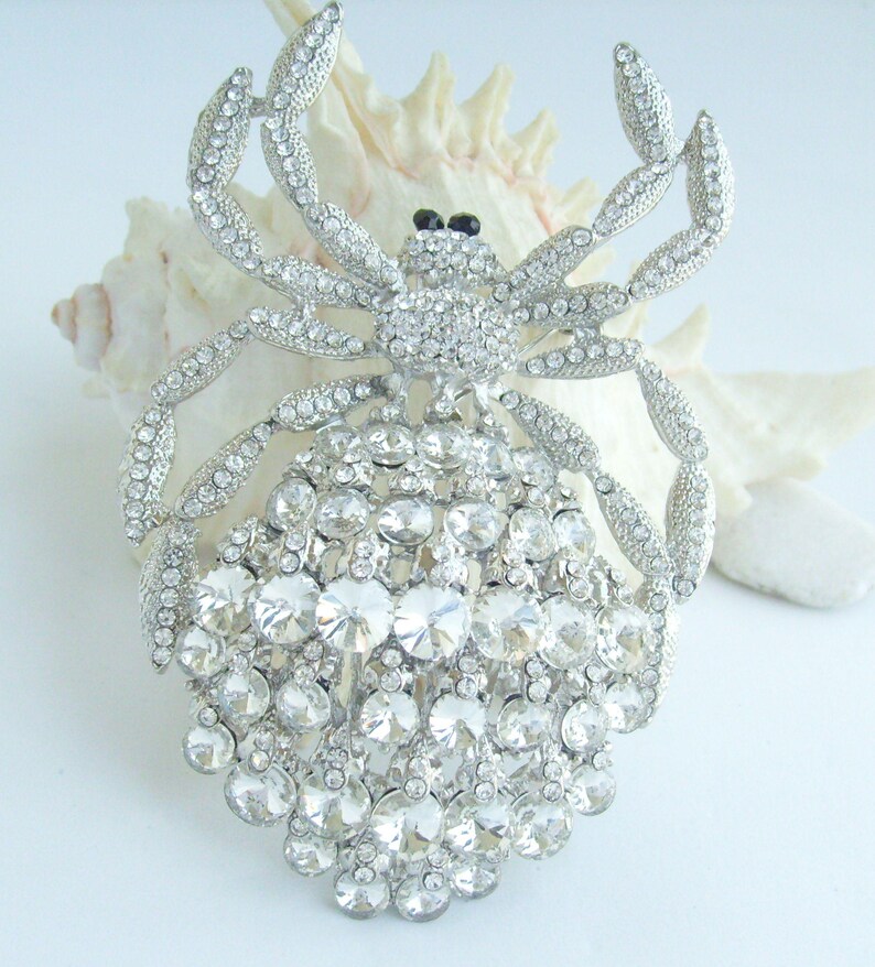 Może przedstawiać: Srebrna broszka w kształcie kraba, inkrustowana przezroczystymi kryształkami. Ciało i nogi kraba są w pełni pokryte błyszczącymi kryształami, z czarnymi akcentami dla oczu. Broszka jest umieszczona na białym tle.