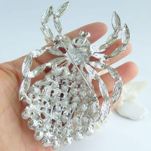 Może przedstawiać: Srebrna broszka w kształcie pająka, ze skomplikowanymi detalami na nogach i ciele. Broszka ma szpilkę z tyłu do bezpiecznego zapięcia. Ciało pająka pokryte jest małymi, okrągłymi ozdobami, dodającymi tekstury i blasku.