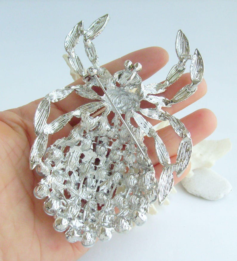 Może przedstawiać: Srebrna broszka w kształcie pająka, ze skomplikowanymi detalami na nogach i ciele. Broszka ma szpilkę z tyłu do bezpiecznego zapięcia. Ciało pająka pokryte jest małymi, okrągłymi ozdobami, dodającymi tekstury i blasku.